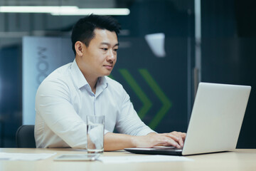 Fototapeta na wymiar Serious and focused young Asian businessman, freelancer, entrepreneur. Sitting at a table in a modern office, working with a laptop and documents.