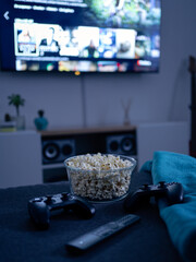 Bowl of popcorn with joysticks, a tv remote control on a sofa in the living room. Selective focus