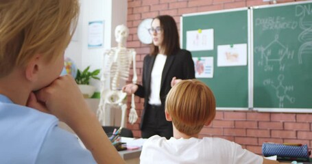 Smart female teacher talking and gesturing while standing near model of human skeleton at classroom. Woman teaching caucasian pupils biology science at modern school. - Powered by Adobe