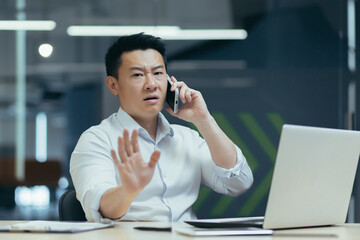 Worried and serious young male Asian businessman talking on the phone in the office. Sitting at a table with a laptop and documents. Trying to solve problems