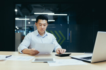 Young Asian male accountant working with documents and calculator. Sitting at a desk with a laptop in a modern office