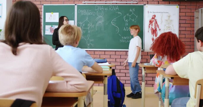 Cute redhead schoolboy standing answering near blackboard at classroom about DNA structure. Female teacher listening answer near, classmates sitting at desks. Biology lesson.