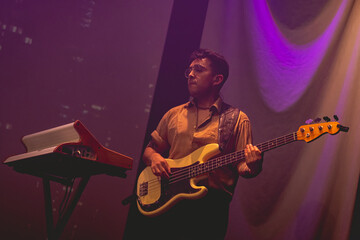 Obraz premium Young latino bassist and keyboardist with shirt and glasses, and cream bass and vintage keyboard playing live in a concert under colorful lights