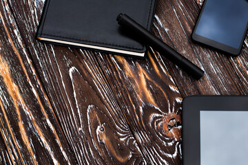 A black tablet, mobile phone and notebook with a leather cover and a pen lie on a dark wooden...