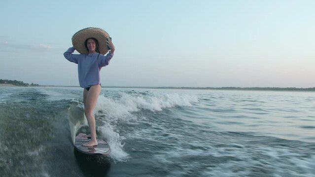 A surfer in a straw hat jumps on a wakeboard and takes a selfie on her phone. An experienced wakeboarder sprays water drops into the camera.
