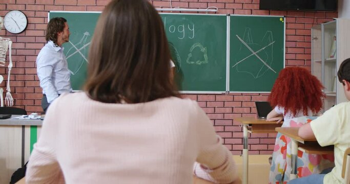 Pretty schoolgirl with brown hair presenting composition on ecology theme while standing at class near blackboard. Male teacher and schoolmates clapping in hands during successful speech.