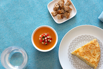 blue tablecloth table. on top a plate of Spanish or potato omelette, gazpacho, croutons, pieces of toasted bread, accompanied by a drink. zenith
