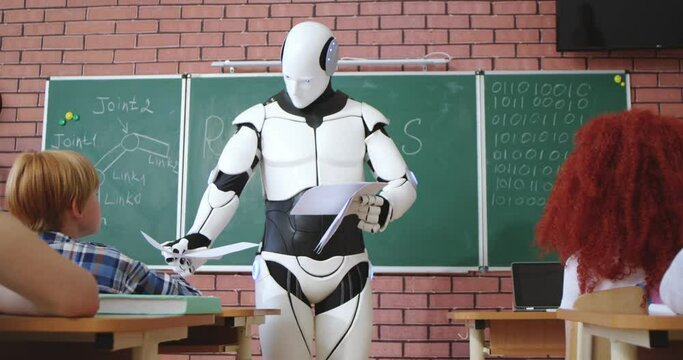 White modern robot walking among desks row while giving elementary school pupils their results of control work. Replacement of human labor by artificial intelligence.