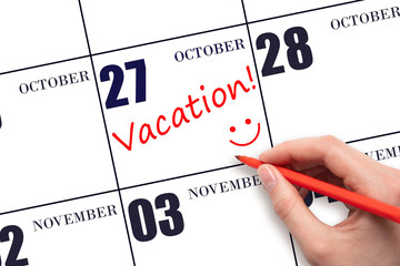A hand writing a VACATION text and drawing a smiling face on a calendar date 27 October. Vacation...