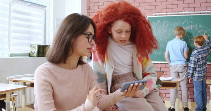 Two female classmates with dark and red hair having chat at classroom during break. Cheerful girls in casual clothes chatting and using smartphone at modern school.