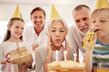 Senior woman celebrating a birthday with family at home, wearing party hats and blowing whistles....