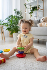 Vertical view of the female kid toddler playing wooden toys at home. Cute little kid sitting on the floor with toy eco-friendly pyramid in natural playroom and having fun