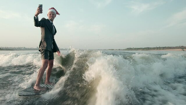 A surfer jumping on a wakeboard in a suit and with a Christmas red hat on his head is photographed on his phone. An experienced wakeboarder sprays water drops into the camera.