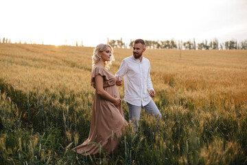 A pregnant woman in a beautiful long dress is walking through the field with her husband in a wheat field at sunset. Future parents. © VikaNorm