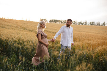 A pregnant woman in a beautiful long dress is walking through the field with husband in a wheat field at sunset. Future parents.