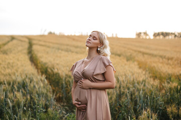 A pregnant blonde woman in a dress is touching her belly in a wheat field at sunset.