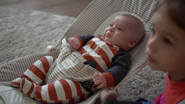 Childhood, motherhood, family, comfort. Little newborn baby boy in striped bodysuit pajamas awake looking around smiles plays rattles sibling sister kid lul amuse brother in room at home atmosphere