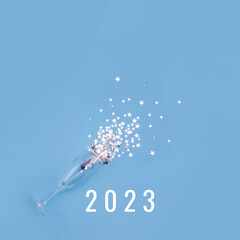 Champagne glass on blue background glass surrounded by silver star confetti and numbers 2023 . New Year celebration or party concept. Selective focus, copy space