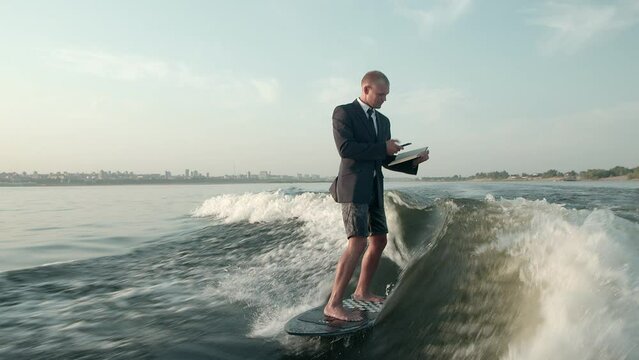 A surfer jumping on a wakeboard in a suit with a book in his hands takes photos of the pages of the book with his phone. An experienced wakeboarder sprays water drops into the camera.