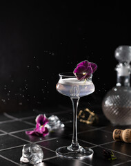 Elegant cocktail with champagne in a crystal glass garnished with purple flower on trendy black tile background with ice cubes.