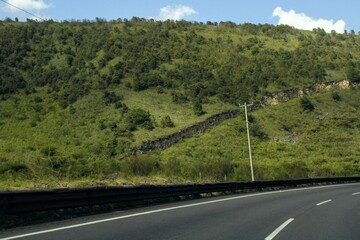 Autopista asfaltada con señalización de carriles y monte de fondo con arboles y arbustos con rocas y un poste de electricidad al fondo 