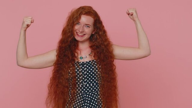 I am strong and independent. Young redhead woman showing biceps and looking confident, feeling power strength to fight for rights, energy to gain success win. Ginger girl on pink studio background