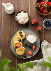 Cottage cheese pancakes with sour cream, milk, blueberries and strawberries on a dark wooden background. Healthy and delicious breakfast. Top view