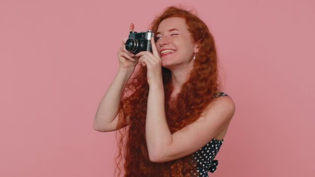 Redhead young woman tourist photographer in black dress and dress taking photos on retro camera and smiling. Travel, summer holidays vacation. Ginger girl indoors isolated on pink studio background