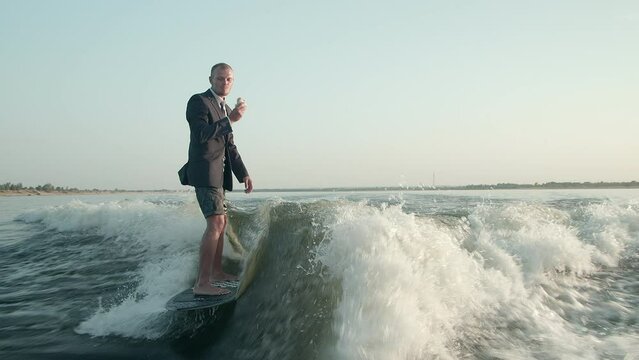 A surfer jumping on a wakeboard in a suit with a bun in his hands. The surfer eats a bun and rides a wakeboard. An experienced wakeboarder sprays water drops into the camera.