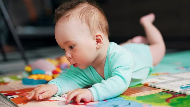 Cute child with funny hairdo reaches the edge of a mat he is lying at. Lovely baby is interested with pictures at the cover.