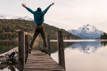 Fototapeta na wymiar Joven saltando de alegria en un muelle de San Carlos de Bariloche. Montañas, lago y paisajes increíbles lo rodean. 