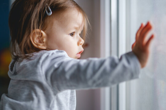 One Small Caucasian Baby Girl Standing By The Window At Home Looking Outside Alone Waiting In Day Side View Copy Space Infant Child Childhood Concept