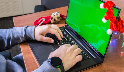 Closeup man hands typing on laptop keyboard with empty green screen and Christmas decoration on background. Holiday concept. Office. Mock-up