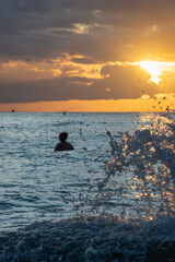 Waves crashing on the shoreline with incredible golden hour sunset in the background and one person swimming in beach.