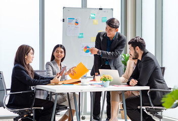 Businessman manager presenter in formal suit standing holding pen pointing at graph chart document on whiteboard presenting company information to Asian male female colleagues in meeting room