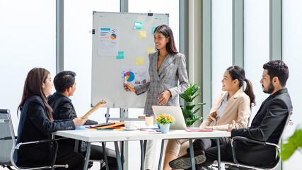 Asian businesswoman presenter in formal suit standing smiling holding pen pointing at graph chart document on whiteboard presenting information to multinational male female colleagues in meeting room