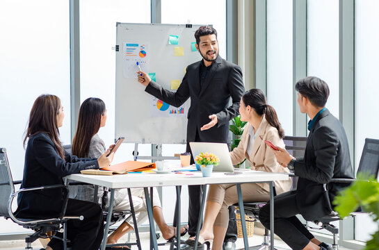 Indian businessman manager presenter in formal suit standing holding pen pointing at graph chart document on whiteboard presenting company information to Asian male female colleagues in meeting room