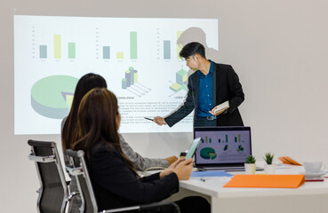 Millennial Asian young cheerful male businessman presenter in formal suit standing holding notebook and pen pointing presenting corporate graph chart data information from wall screen in meeting room