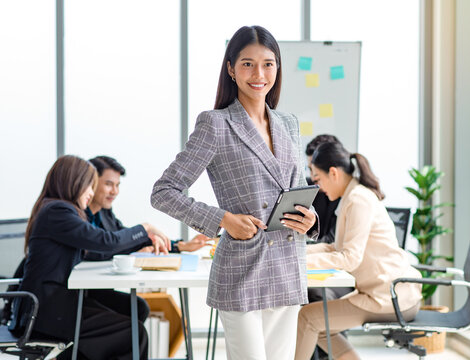 Portrait shot of millennial Asian cheerful successful professional businesswoman entrepreneur in formal suit standing smiling crossed arms in meeting room while employee and colleague brainstorming
