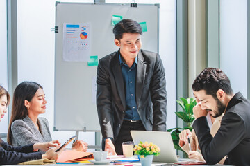 Businessman manager presenter in formal suit standing holding pen pointing at graph chart document on whiteboard presenting company information to Asian male female colleagues in meeting room