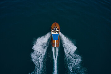 Wooden expensive italian boat with people fast movement on the water top view. High speed open modern wooden boat moving fast on dark water aerial view. - obrazy, fototapety, plakaty