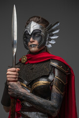 Fototapeta premium Shot of ancient valkyrie with make up dressed in dark armor with red cape holding spear.
