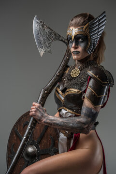 Portrait of wild valkyrie dressed in armor holding huge axe and looking at camera against grey background.