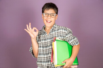 Happy Latin ten year old student in eyeglasses holding folders and papers while doing an OK gesture standing on purple background. Back to school concept.