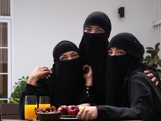 Three pretty Muslim women wearing black hijab or niqab sitting outdoors having dates, apples and juice looking at camera.