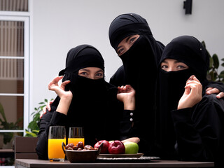 Three pretty Muslim women wearing black hijab or niqab sitting outdoors having dates, apples and juice looking at camera.