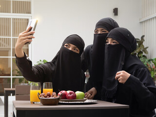 Portrait of three Muslim women wearing black hijab or niqab taking selfie using smartphone while sitting together outdoors at cafe with dates and apples on the table.