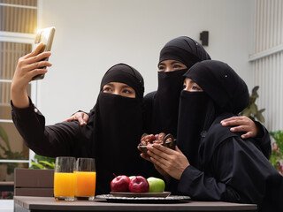 Portrait of three Muslim women wearing black hijab or niqab taking selfie using smartphone showing bowl full of dates while sitting together outdoors at cafe.