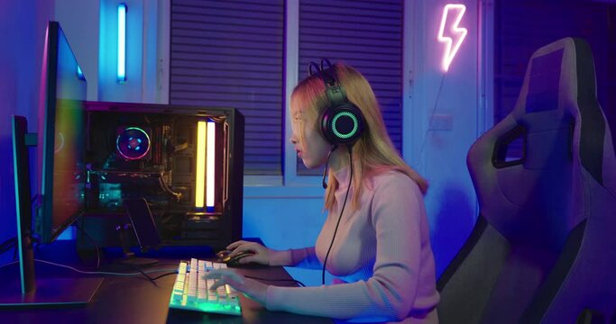 Asian professional gamer playing online video game on desktop computer PC have colorful neon LED lights, young woman in gaming headphones using computer for playing game at home