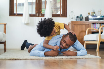Black family have fun playing together on floor at home. Laughing African American boy carrying on Dad's back and tickling in living room at home. Black father play together with son after work. - obrazy, fototapety, plakaty